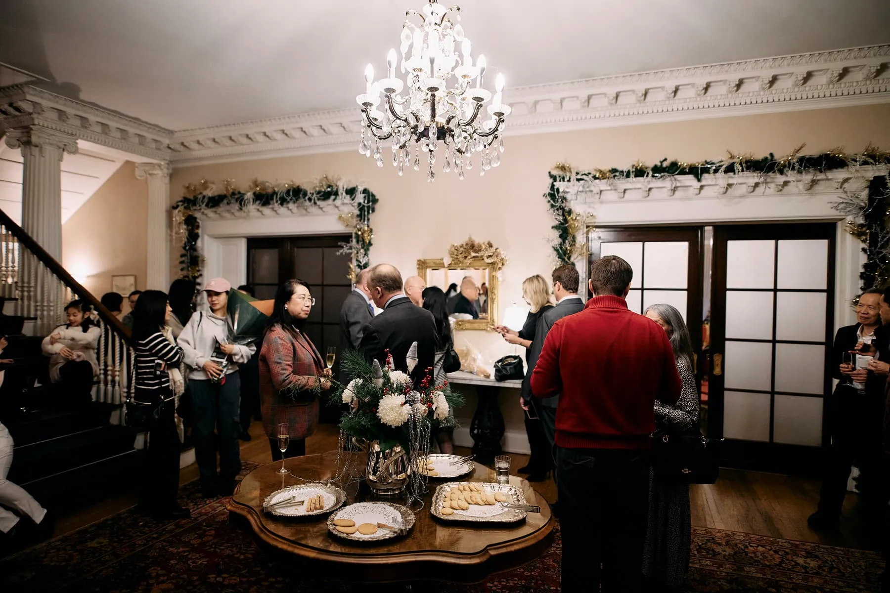 Wide view of a decorated reception room with a chandelier, holiday garland and guests gathered around a table, at a Vancouver private event.