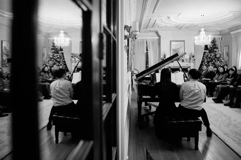 Black and white photograph of two pianists at two grand pianos, reflected in a mirror beside a Christmas tree.