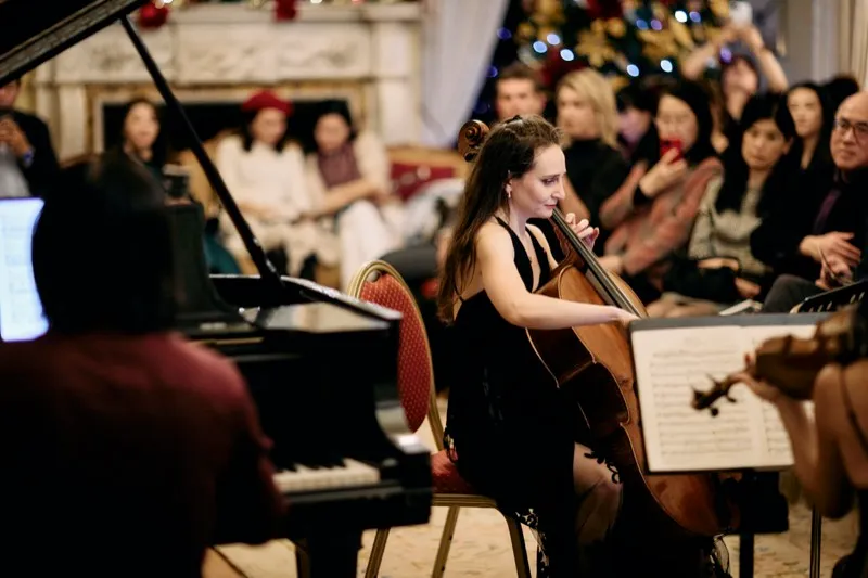 A cellist performs beside a grand piano in front of a Christmas tree, the audience watching.