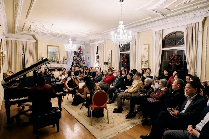 A wide view of a chamber concert in a period drawing room, the audience seated around a grand piano.