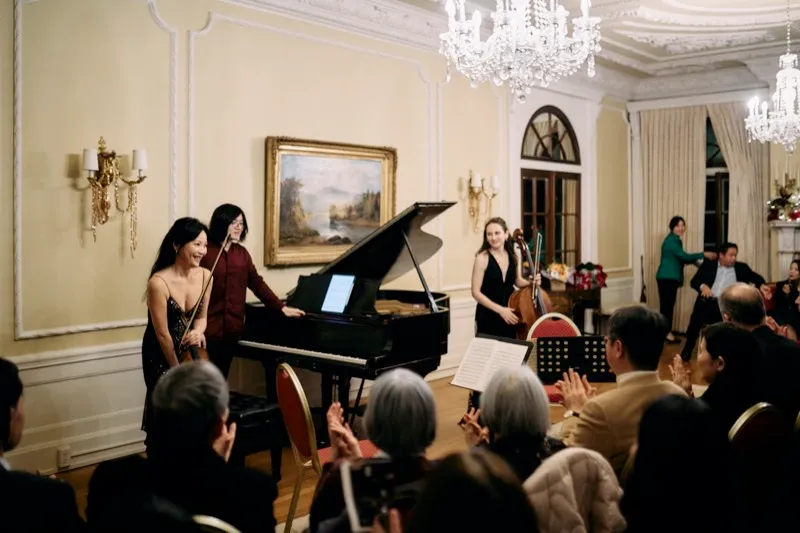 Performers and audience under chandeliers in a cream drawing room, the grand piano at the centre.