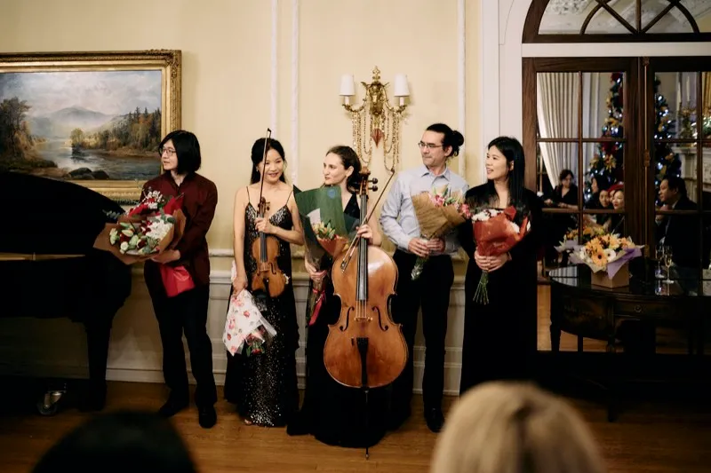 Five musicians hold bouquets beside a grand piano after a recital.