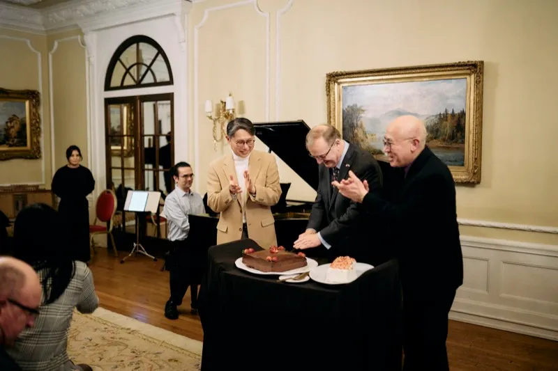 Three hosts applaud beside a chocolate cake on a black-draped table at the end of a recital.