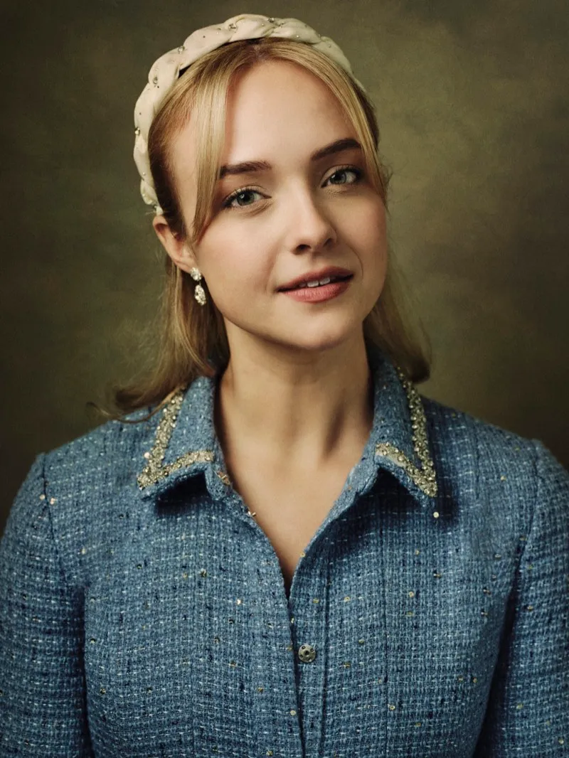 Headshot of a blonde woman in a pale blue tweed jacket with a pearl-trimmed collar and a padded headband, on a dark olive studio backdrop.