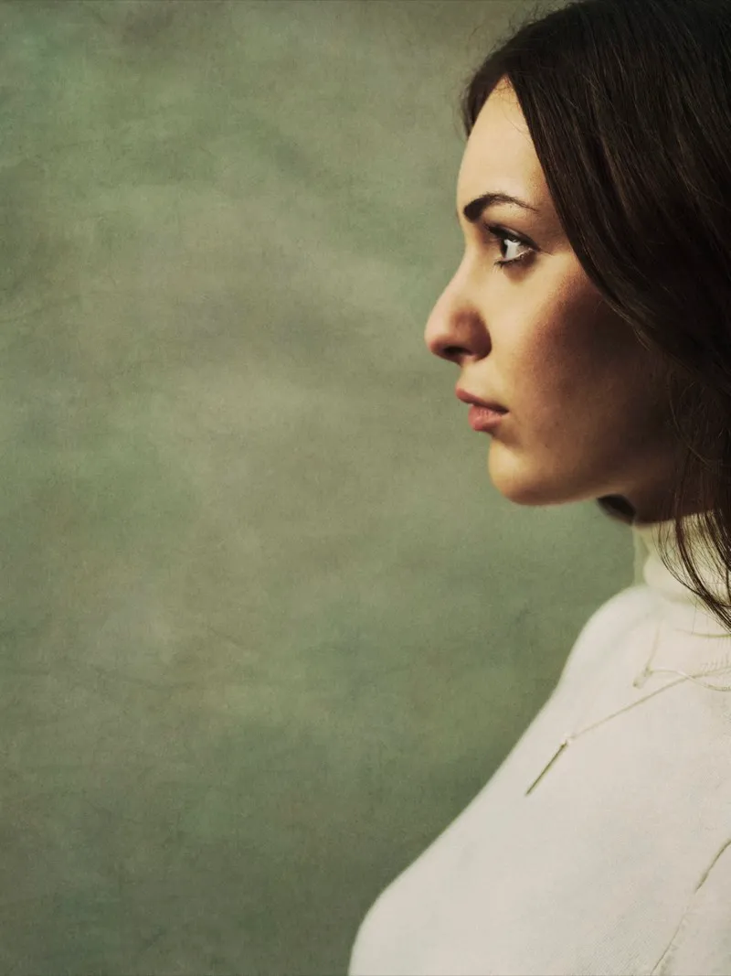 Profile portrait of a dark-haired woman in a cream turtleneck, looking to the left against a soft green painted backdrop.