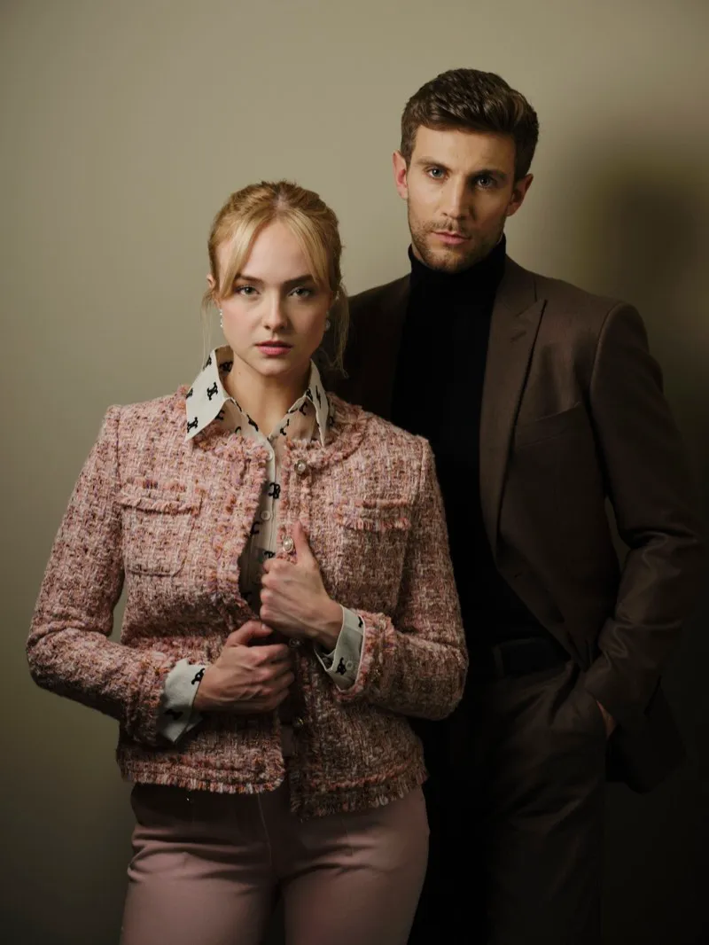 Studio portrait of a couple: a blonde woman in a pink tweed jacket in front, a man in a brown suit and black turtleneck behind her.