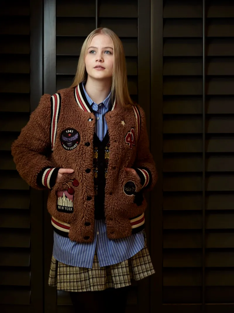 Full-length studio portrait of a young blonde woman in a brown varsity jacket, striped shirt and plaid skirt, in front of dark window shutters.