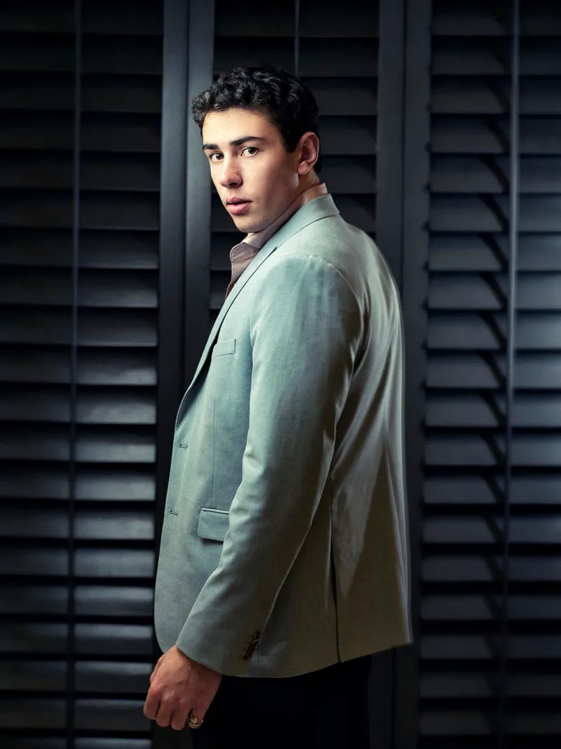 Three-quarter portrait of a dark-haired man in a pale blue suit, looking back over his shoulder in front of black shutters.