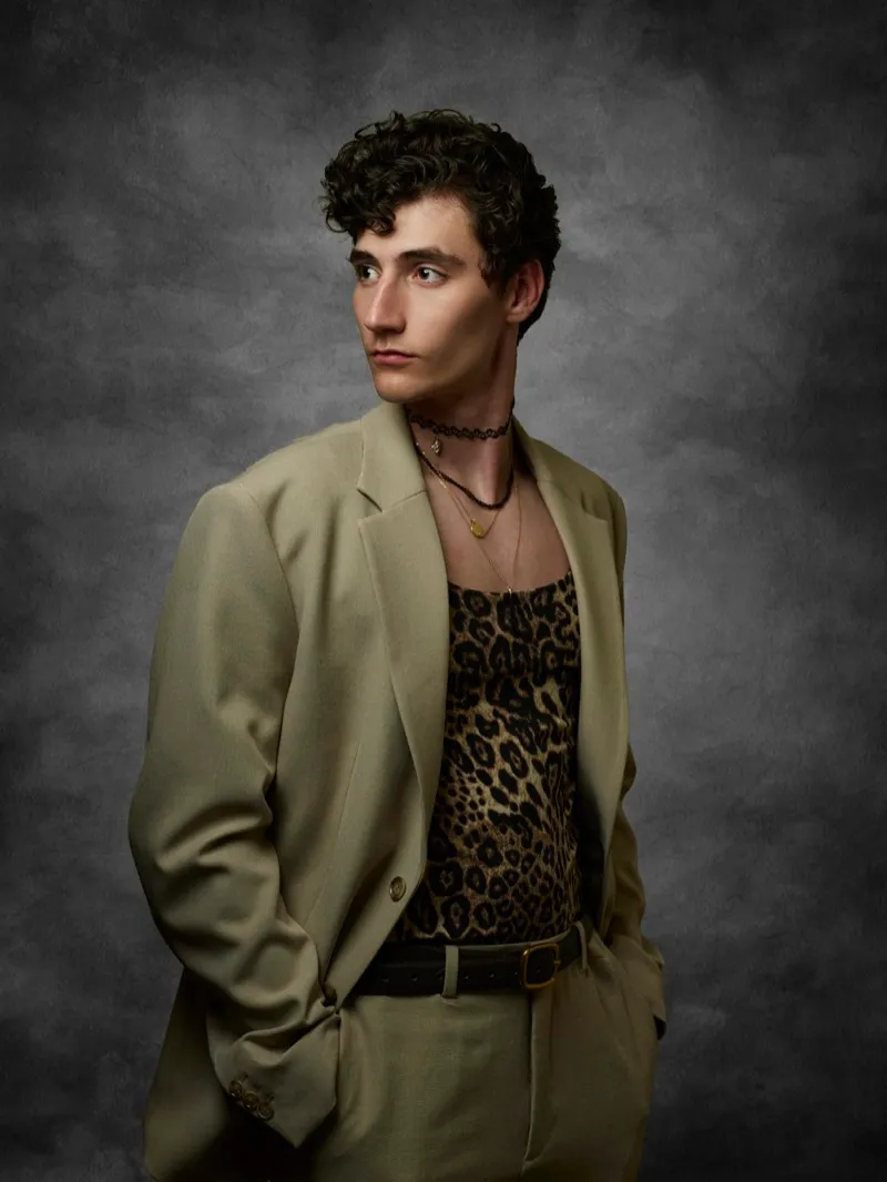 Portrait of a young man with curly dark hair in an oversized khaki suit over a leopard-print top, looking off to the side against a grey studio backdrop.