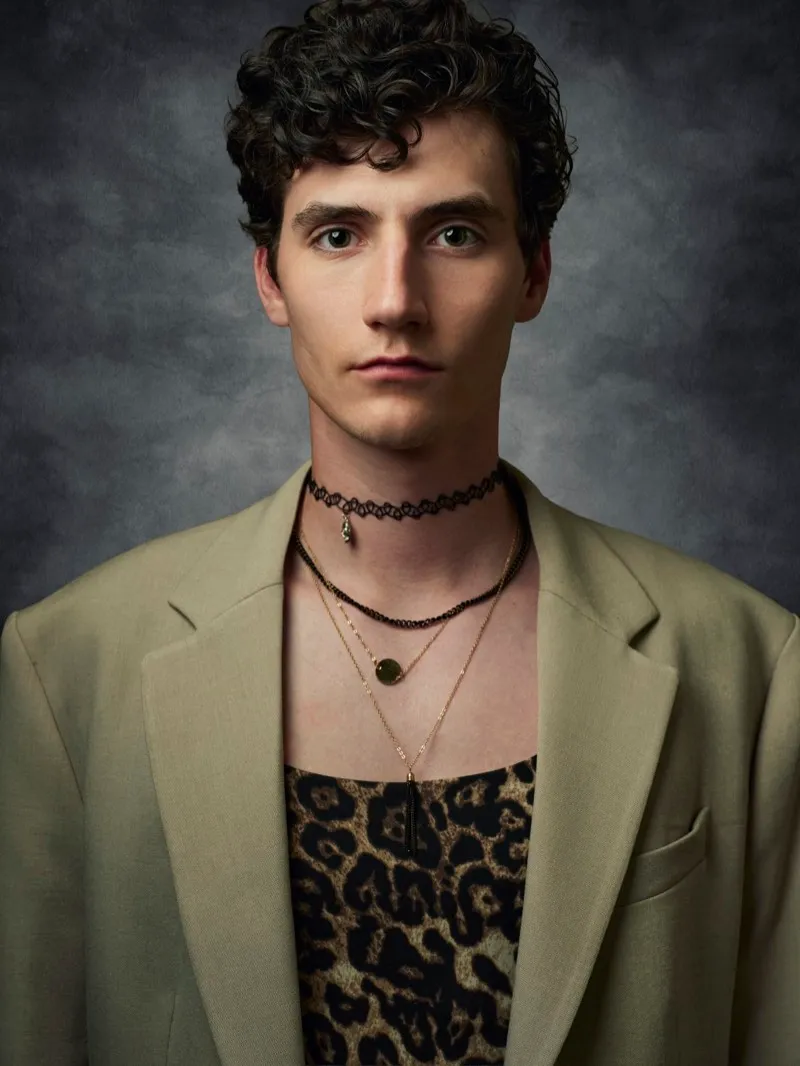 Headshot of a young man with curly dark hair, a choker and layered necklaces, in a khaki jacket over a leopard-print top on a grey backdrop.
