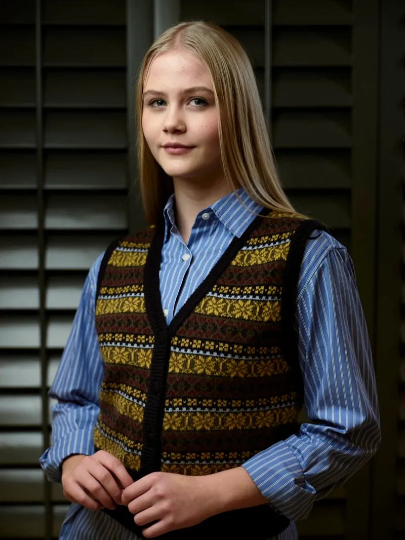 Studio portrait of a young blonde woman in a patterned knitted vest over a blue striped shirt, standing in front of dark shutters.