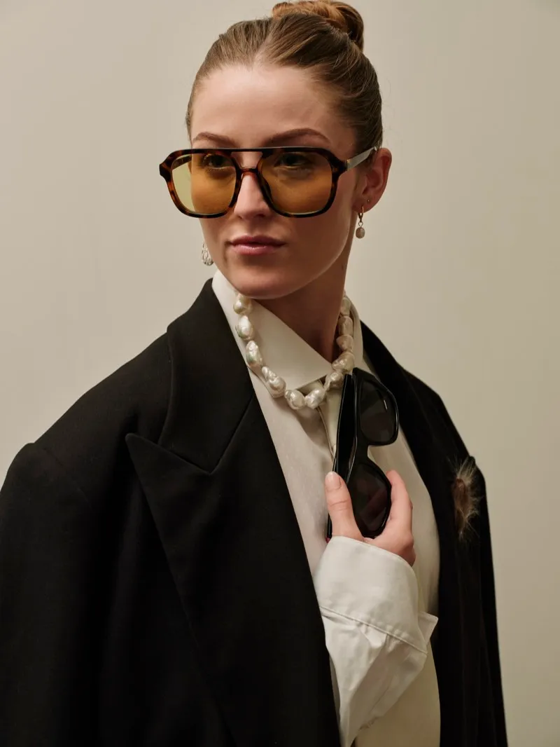 Editorial headshot of a woman with red hair pulled back, in amber-tinted glasses, a large pearl necklace and a black jacket over a white shirt.