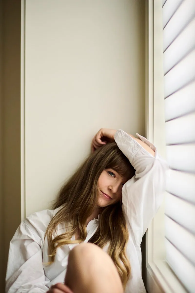 Window-light portrait of a woman in a white shirt sitting on a windowsill with one arm resting on her head, smiling softly.