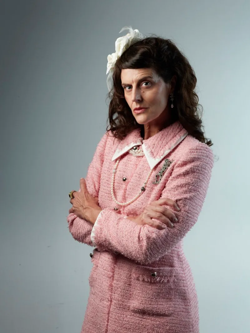 Studio portrait of a woman in a pink tweed suit with a white flower in her hair, arms crossed, against a pale blue backdrop.