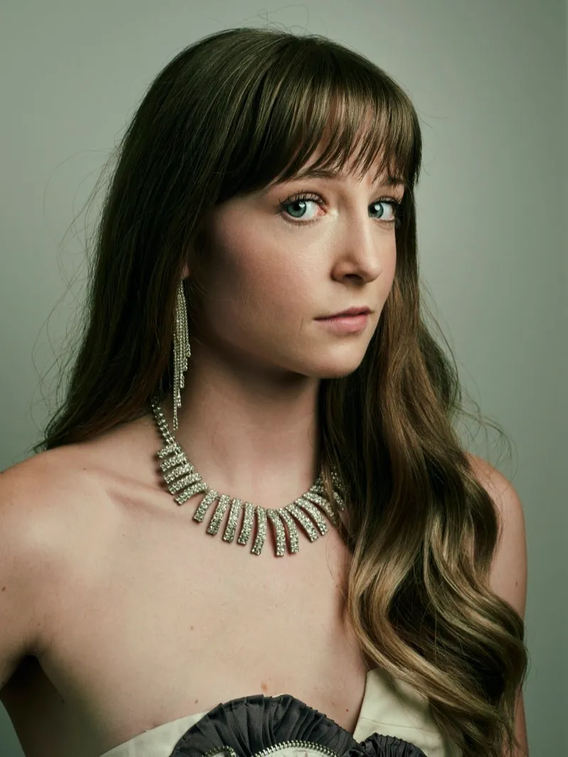 Headshot of a woman with long brown hair and a fringe, wearing a crystal statement necklace and drop earrings, on a sage backdrop.