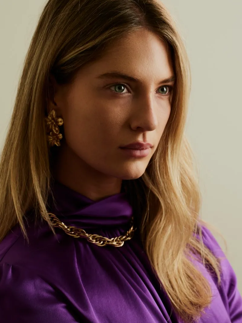 Headshot of a blonde woman in a purple satin top with a chunky gold chain and gold earrings, looking to the side on a cream backdrop.