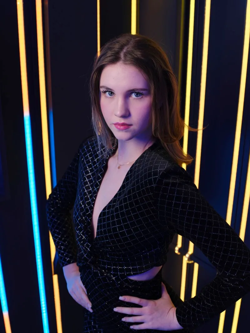 Portrait of a young woman in a black sequinned jumpsuit, hands on her hips, framed by vertical blue and yellow light strips.
