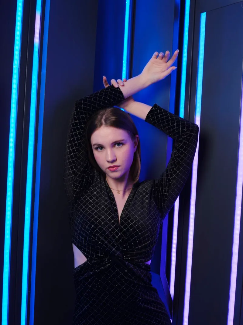 Portrait of a young woman in a black sequinned outfit with her arms raised above her head, between vertical blue and yellow light strips.