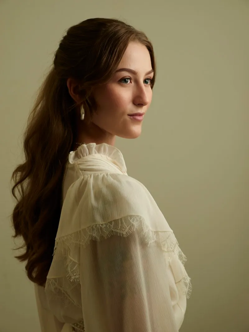 Three-quarter headshot of a young woman with long brown hair in a cream ruffled blouse, looking to the side on a warm beige backdrop.