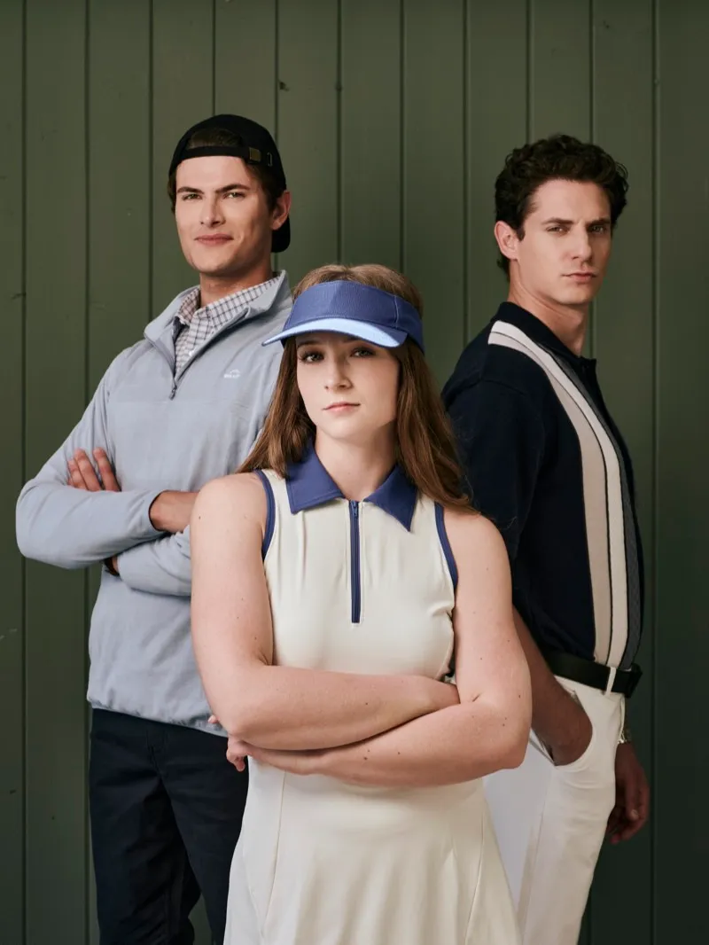 Studio portrait of a woman in a white tennis dress and visor with her arms crossed, flanked by two men in sportswear, against green panelling.