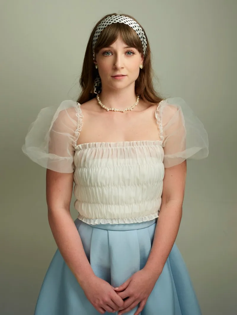 Studio portrait of a woman in a cream puff-sleeved blouse, pale blue skirt and patterned headband, hands clasped, on a soft green backdrop.