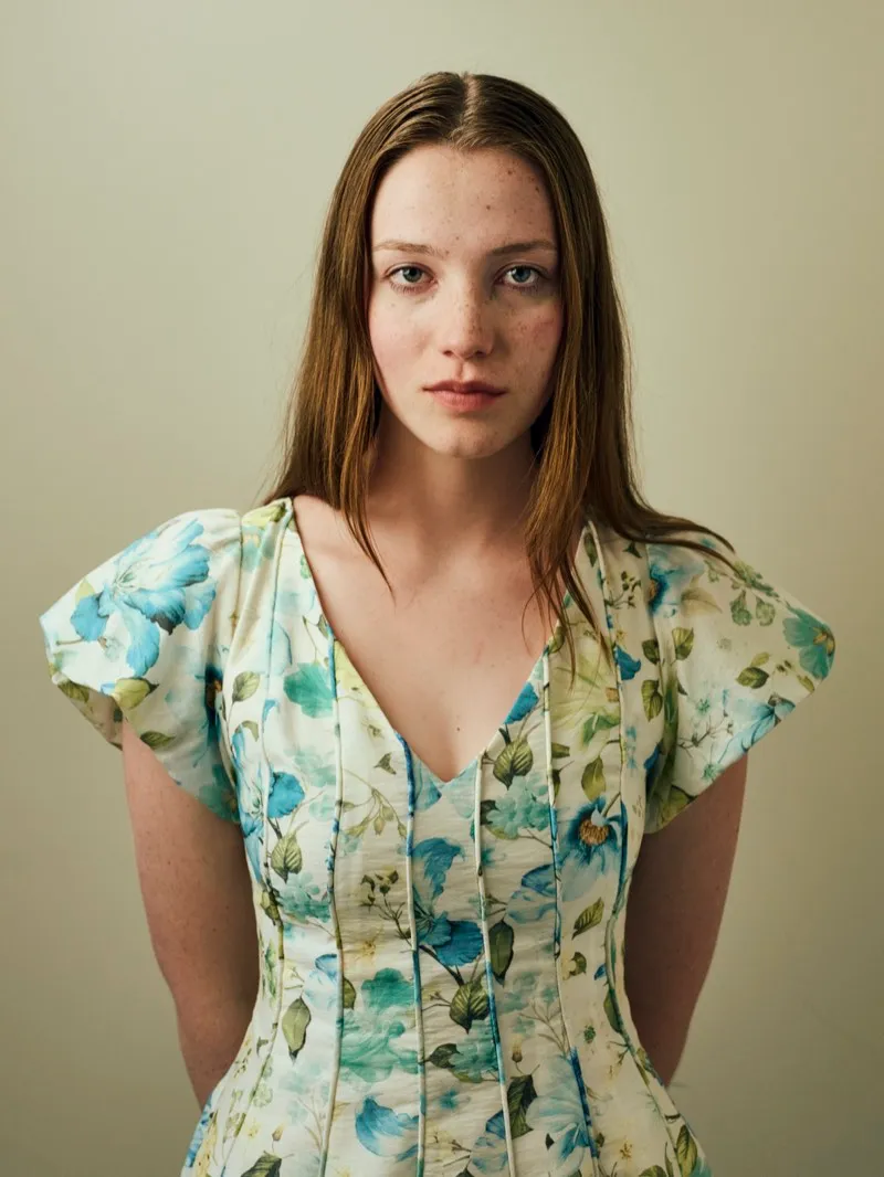 Headshot of a young woman with long straight hair and freckles, in a blue and green floral dress, on a cream studio backdrop.