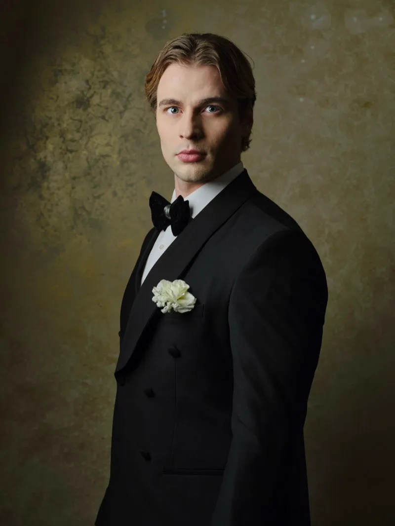 Studio portrait of a fair-haired man in a black tuxedo with a bow tie and a white flower on his lapel, against a gold mottled backdrop.