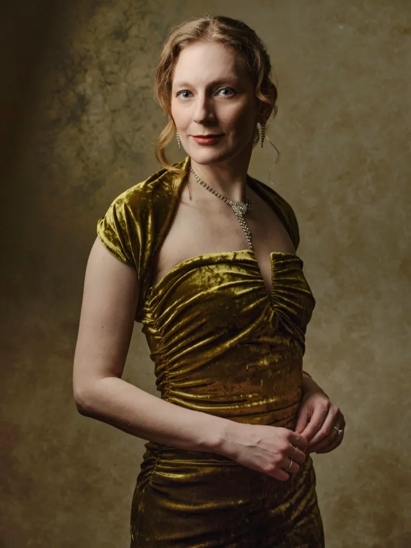 Studio portrait of a fair-haired woman in a gold crushed-velvet dress and a fine chain necklace, against a mottled gold backdrop.