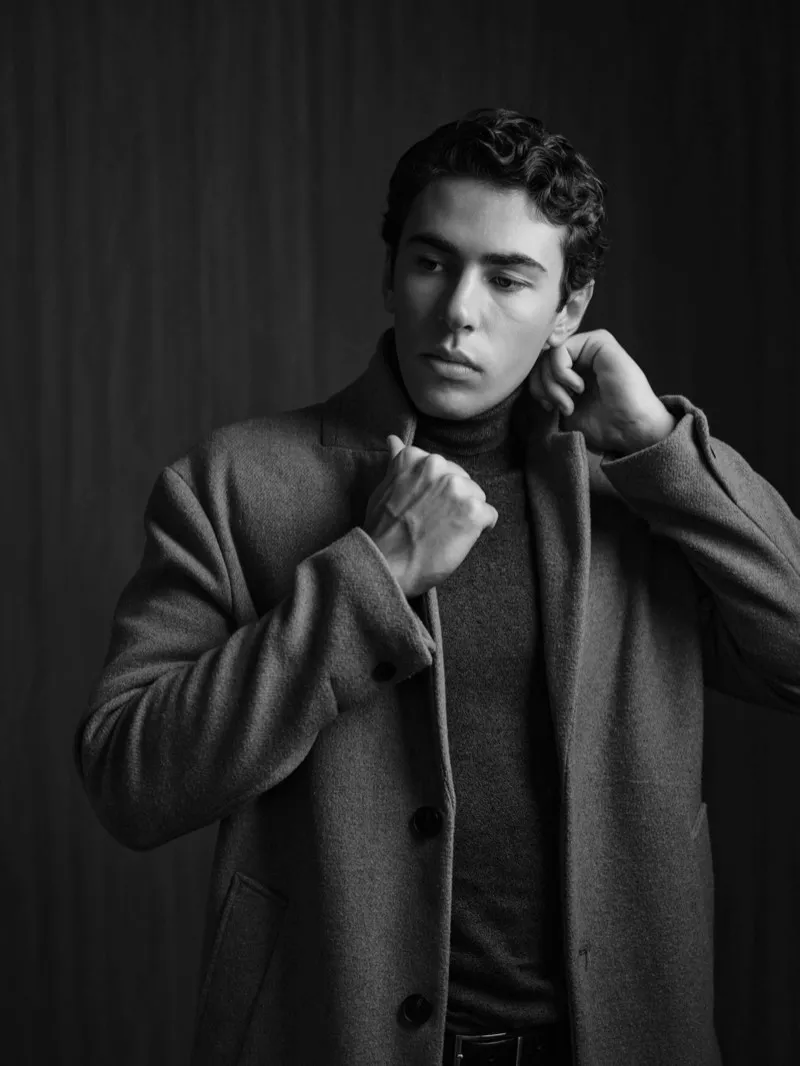 Black and white portrait of a young man with dark curly hair turning up the collar of a wool overcoat over a turtleneck.