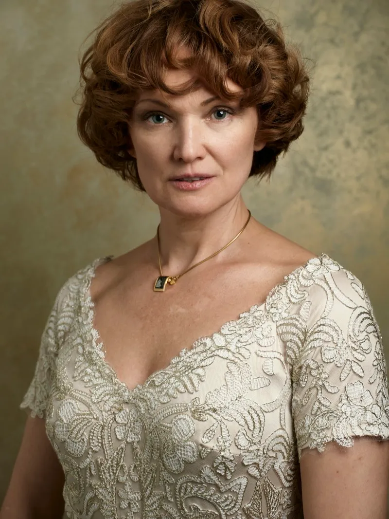Headshot of a woman with short auburn curls in an embroidered ivory dress and a gold pendant, against a mottled gold backdrop.