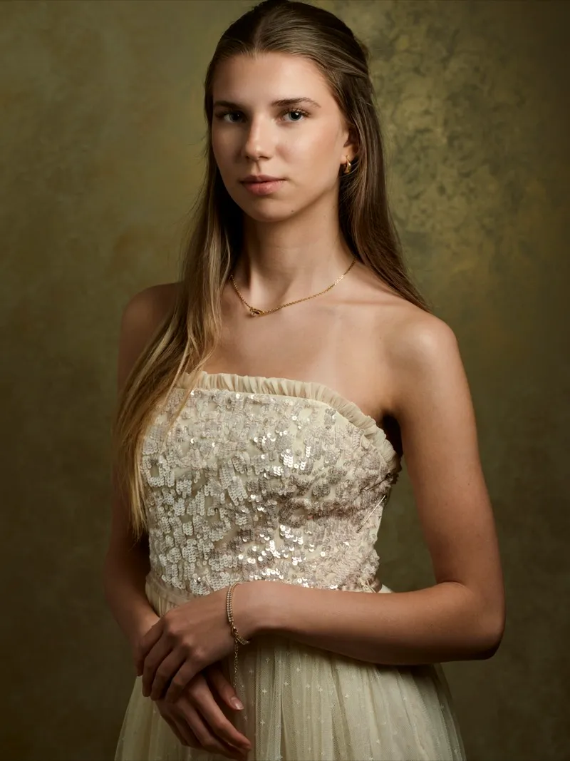 Studio portrait of a young woman with long hair in a sequinned strapless gown, hands clasped, against a gold mottled backdrop.