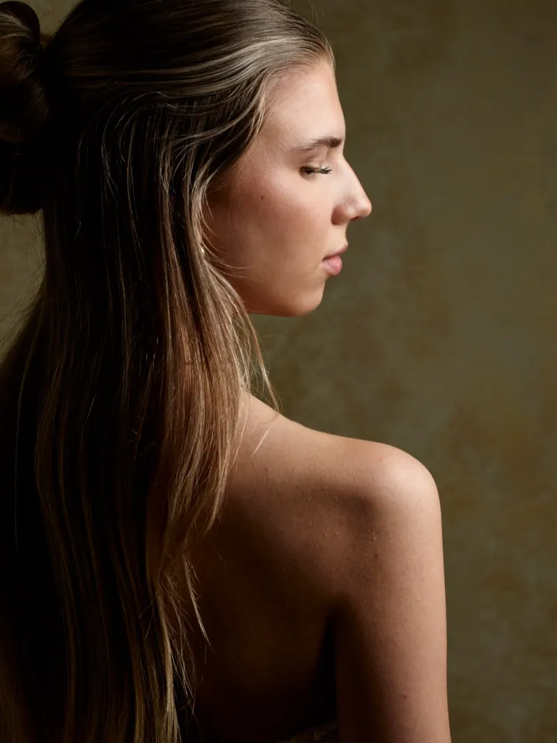 Profile portrait of a young woman with her hair pulled back, bare shoulders turned to the camera, on a warm beige backdrop.