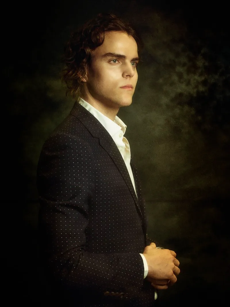 Three-quarter portrait of a man with auburn curls in a dark pin-dot suit and an open white shirt, lit from one side against a dark backdrop.