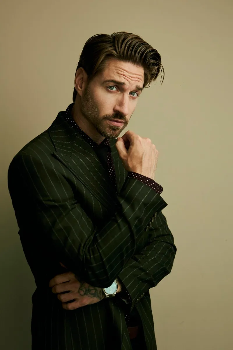 Portrait of a bearded man in a green pinstripe suit with one eyebrow raised and a hand at his chin, on a warm studio backdrop.