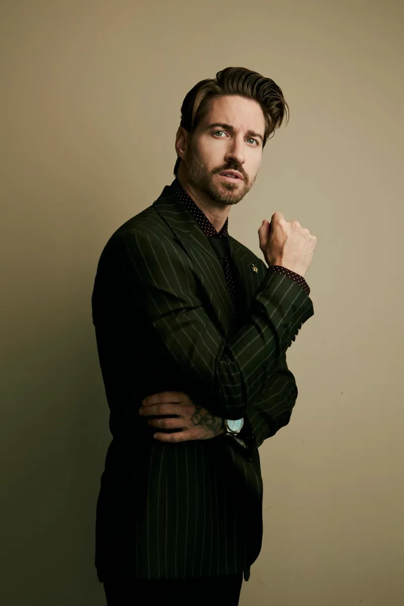Three-quarter portrait of a bearded man in a green pinstripe suit, arms folded, glancing to the side on a warm backdrop.