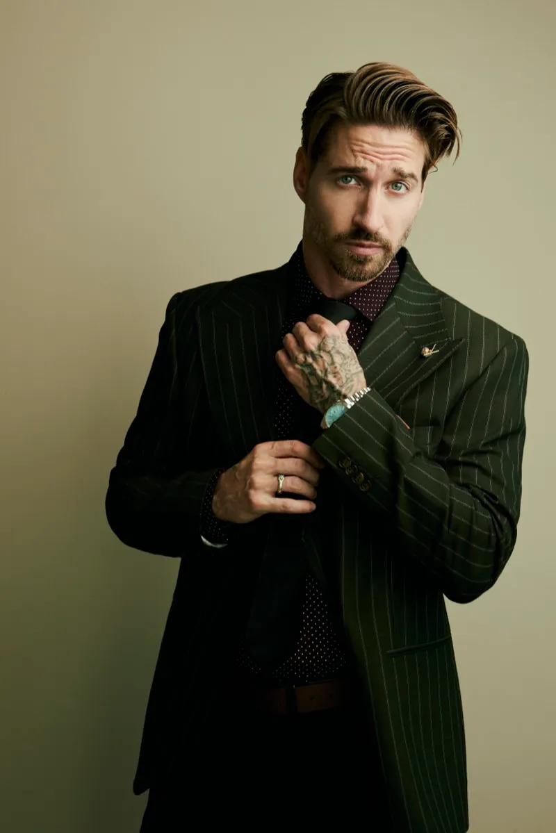 Portrait of a bearded man in a green pinstripe suit with tattooed hands clasped at his chest, on a warm backdrop.