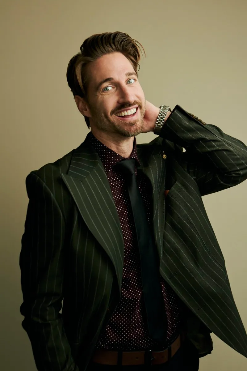 Portrait of a bearded man in a green pinstripe suit and dark tie, smiling with a hand behind his neck, on a warm backdrop.