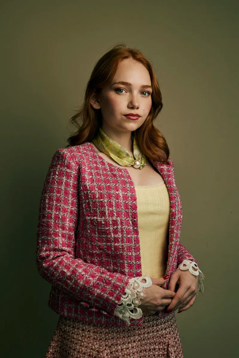 Studio portrait of a red-haired woman in a pink tweed jacket over a yellow top with a patterned silk scarf, hands together.