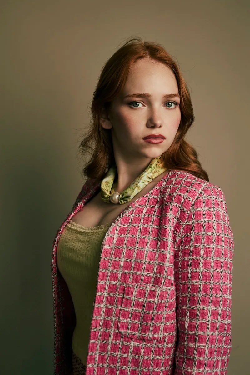 Headshot of a red-haired woman in a pink checked tweed jacket and green scarf, looking straight into the camera on a warm backdrop.