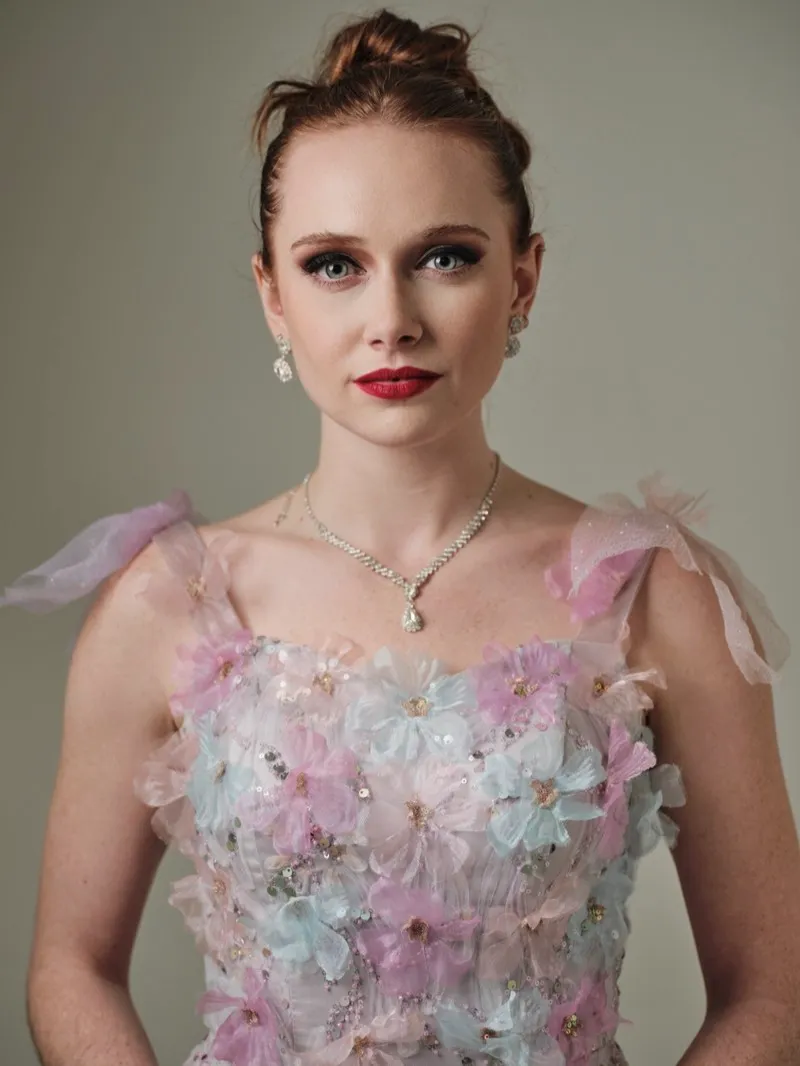 Headshot of a red-haired woman with an updo, red lipstick and drop earrings, in a pastel floral bodice on a pale grey backdrop.