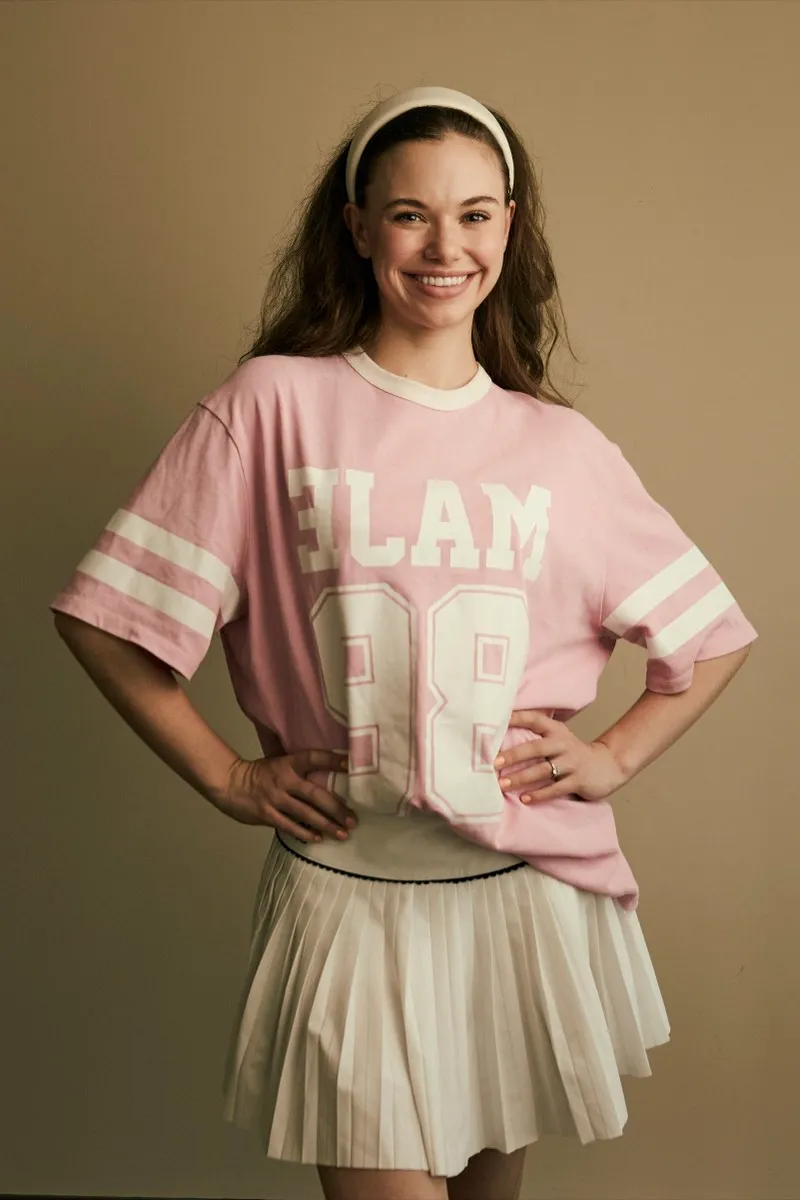 Full-length portrait of a smiling woman in a pink varsity tee, white pleated skirt and headband, hands on her hips, on a beige backdrop.