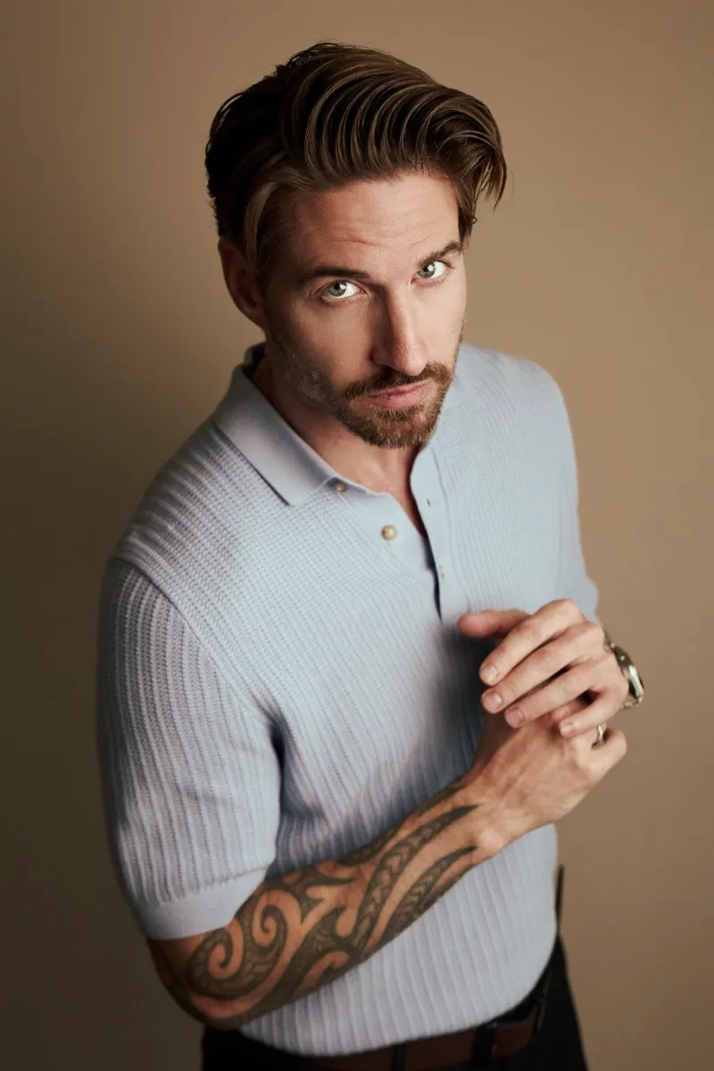 Headshot of a bearded man with slicked-back hair in a pale blue knitted polo, tattooed forearm crossed, on a warm studio backdrop.