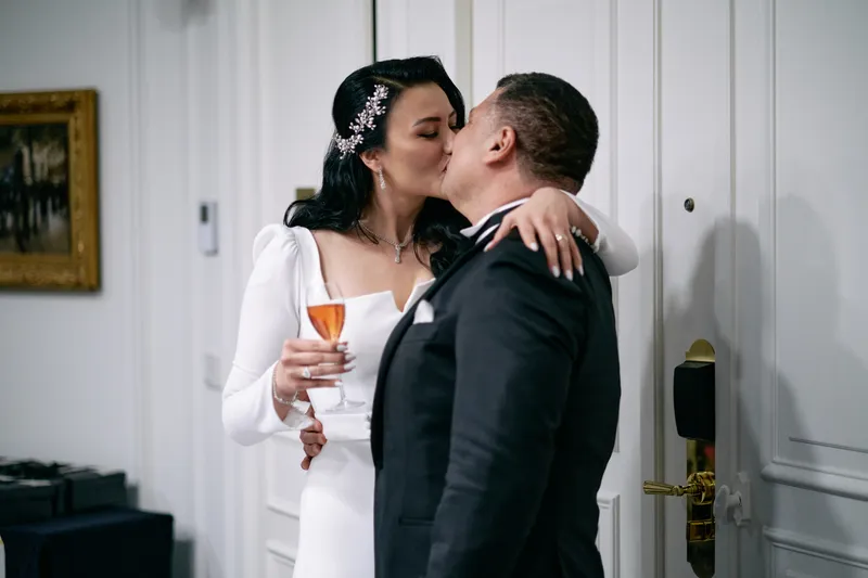 Michael and Julie kiss, each still holding a glass, beside the red roses.