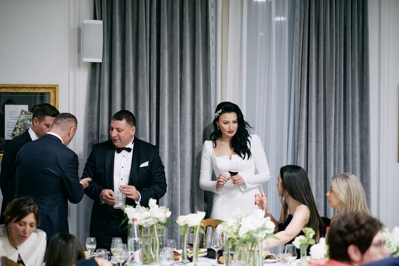 A guest in a dark jacket leans across to speak with the bride at the table.