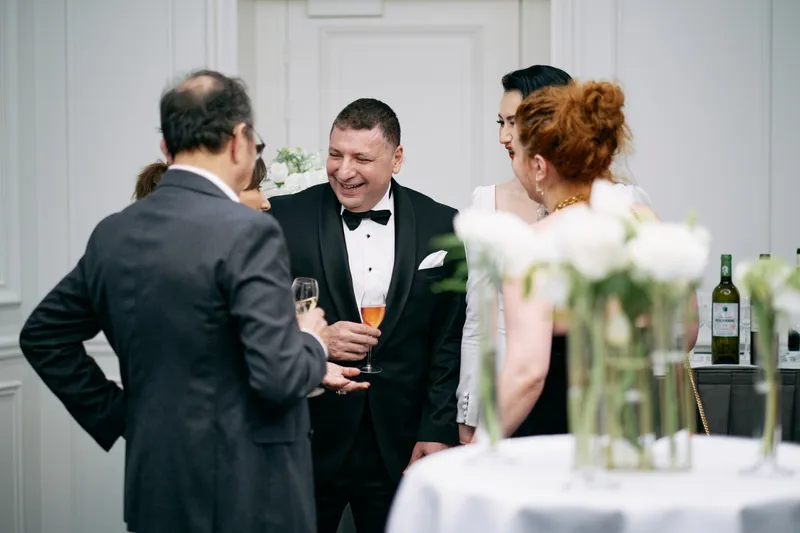 The groom shakes hands with an arriving guest while others gather by the white flowers.