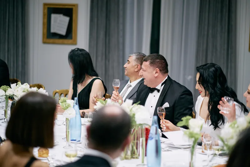 A guest in a black tuxedo laughs with the woman beside him at the dinner table.