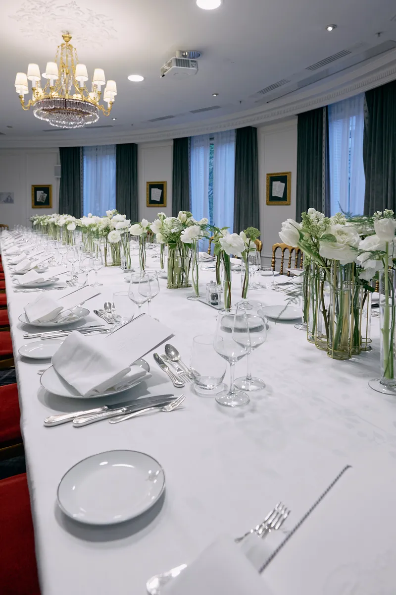 A place setting with folded napkin and silver cutlery, white flowers in glass vases down the table and tall windows behind.