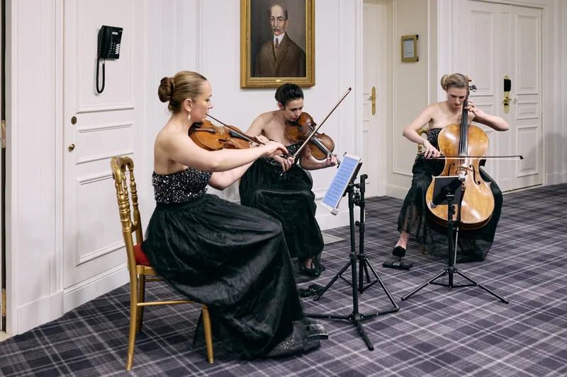 The string trio plays on in their gold chairs beneath the framed portrait while dinner continues.