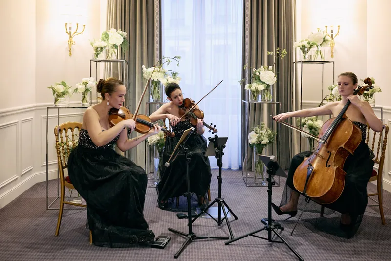 Two violinists and a cellist in black dresses play on gold chairs beside tall white flower stands.