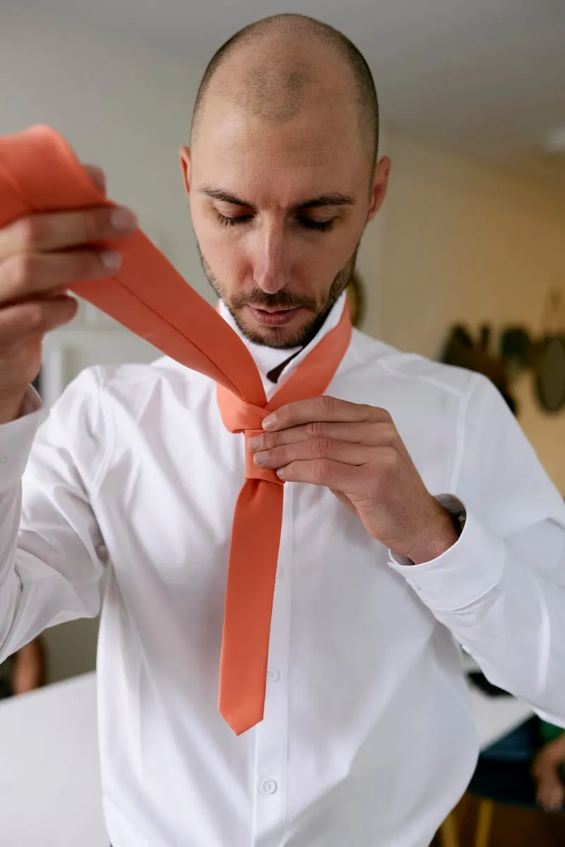 James knotting a coral tie in a white shirt.